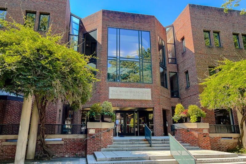 The entrance to The Wharton School, a red-brick building with large glass windows and greenery under a clear blue sky, welcomes visitors—an iconic stop on any Northeast College Tour. Steps lead up to the front doors.