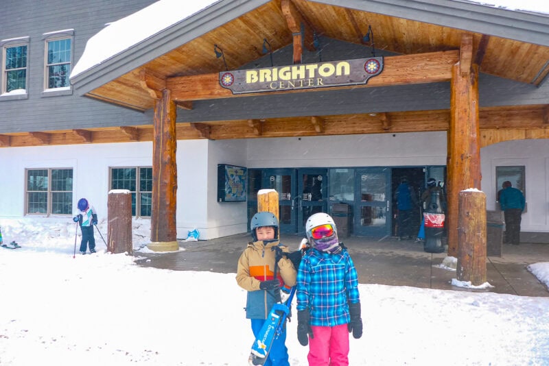 Two children in winter gear stand holding snowboards in front of the Brighton Center ski lodge at this popular Utah ski destination, with snow on the ground and people in the background near the entrance.