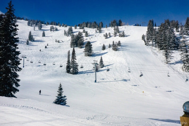 A snow-covered Utah ski slope with scattered pine trees, a few skiers, and a ski lift carrying people up the mountain under a clear blue sky.