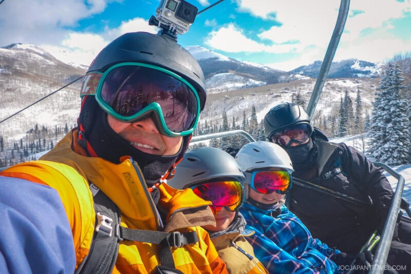 A group of four people wearing ski helmets and goggles ride a Utah Ski lift over snowy mountains and trees, smiling in winter gear under a partly cloudy sky.