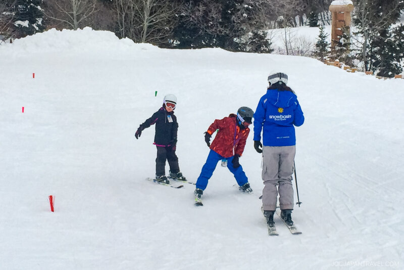 A Utah Ski instructor in blue and gray helps two young children in colorful snowsuits and helmets practice skiing on a snowy slope lined with small marker poles.
