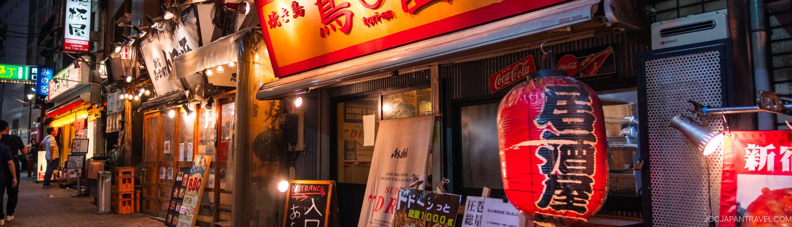 A lively Japanese street at dusk lined with izakayas and restaurants, featuring bright lanterns, illuminated signs, and menu boards, with a few people walking in the distance.