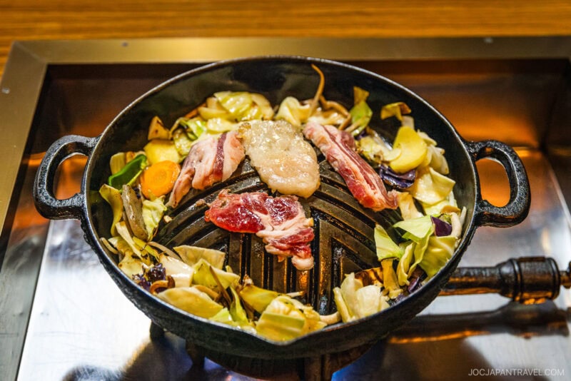 A round cast iron grill pan filled with vegetables and yamagata beef slices cooking on top, with a piece of fat in the center, ready to be grilled at a restaurant table.