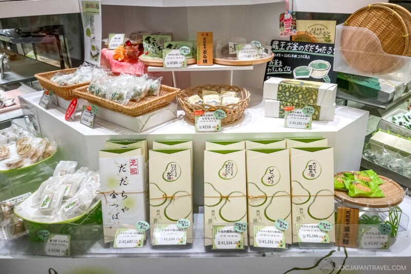 A display of neatly arranged Japanese sweets and matcha products from Yamagata in a shop, with various green and white packaging and price tags in Japanese, along with baskets holding individually wrapped treats.