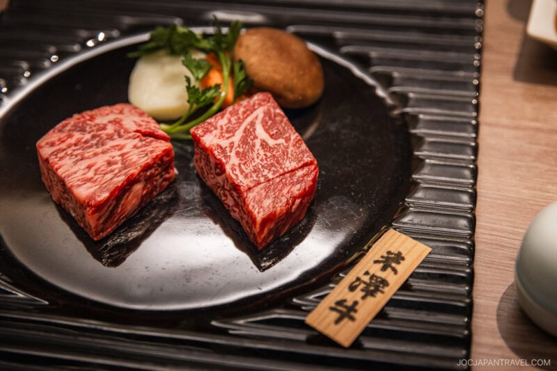 Two thick, marbled slices of Yamagata Wagyu beef are displayed on a black plate with a piece of shiitake mushroom, onion, and green garnish, alongside a wooden label with Japanese characters.