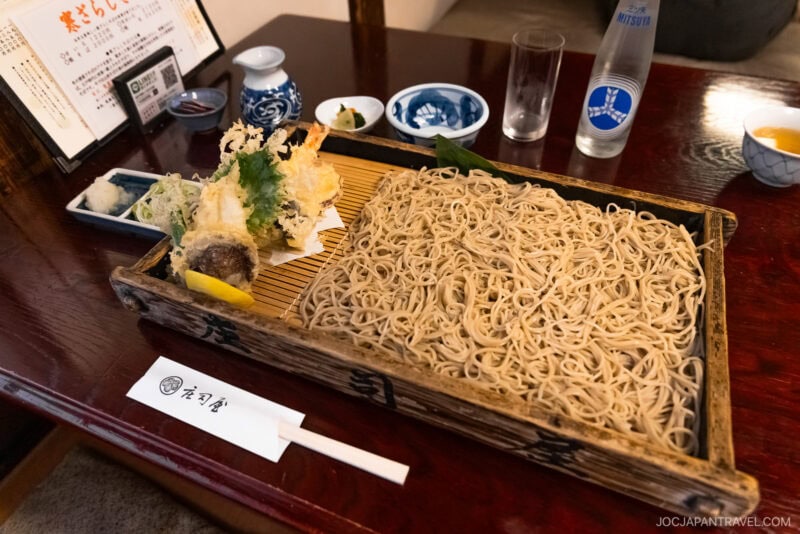 A tray of cold soba noodles, a Yamagata specialty, with tempura vegetables and shrimp, served with dipping sauce, chopsticks, and condiments on a wooden table in a traditional Japanese restaurant setting.
