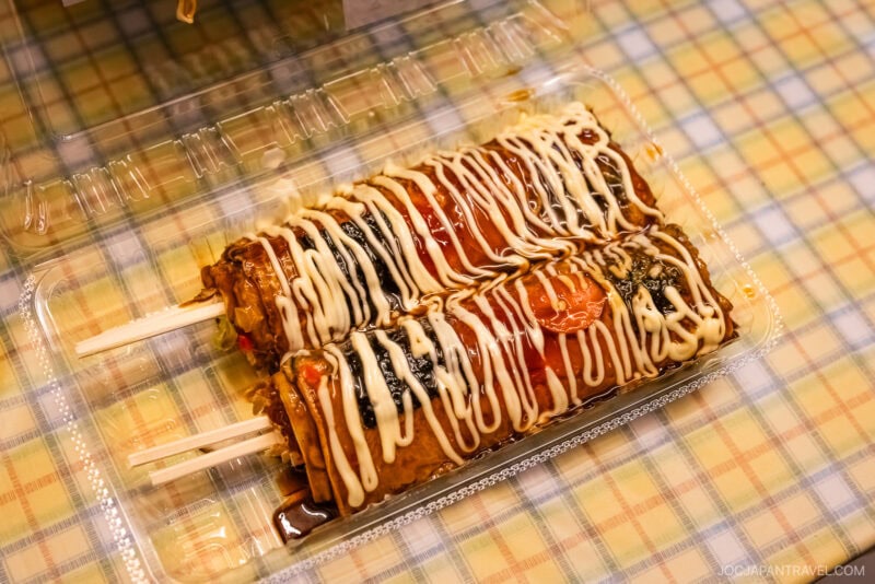 Two Japanese savory skewers, inspired by Yamagata flavors, possibly okonomiyaki on sticks, are topped with zigzag lines of mayonnaise and savory sauce in a clear plastic container on a plaid tablecloth.