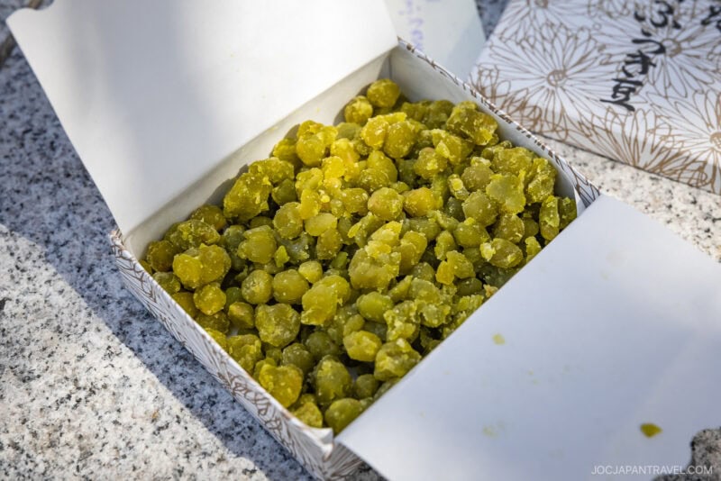 An open box filled with small, round, yellow-green sugared beans from Yamagata sits on a speckled stone surface. The box features a floral patterned design and Japanese writing on the inside flap.