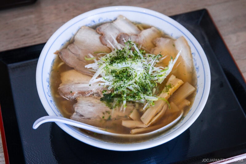 A bowl of Yamagata ramen with sliced pork, bamboo shoots, shredded green onions, and herbs in clear broth, served with a white spoon on a black tray.