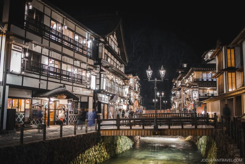 A picturesque night scene of Ginzan Onsen in Yamagata, Japan, featuring traditional wooden ryokan inns illuminated along a narrow canal with a small wooden bridge and ornate street lamps.