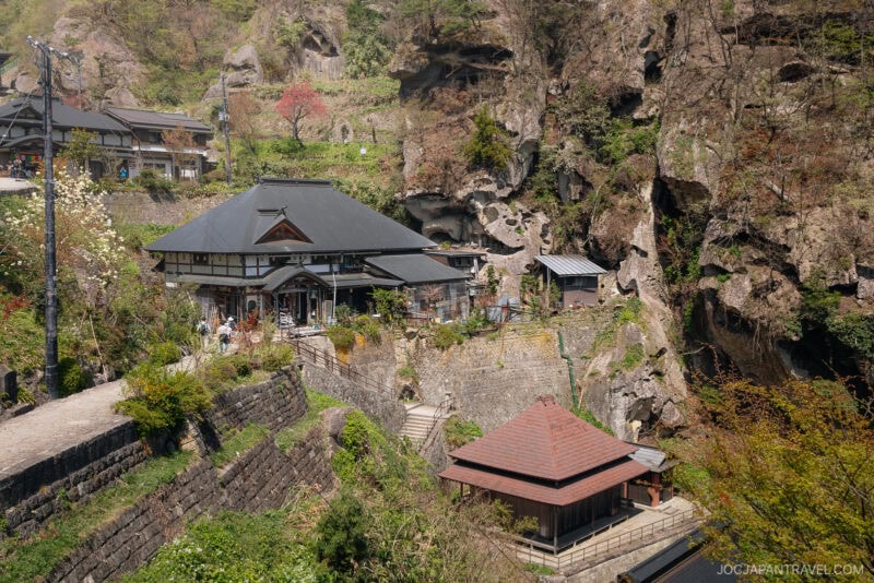 A traditional Japanese building in Yamagata is surrounded by lush greenery and rocky cliffs, with stone steps and smaller structures nestled in the landscape on a sunny day.