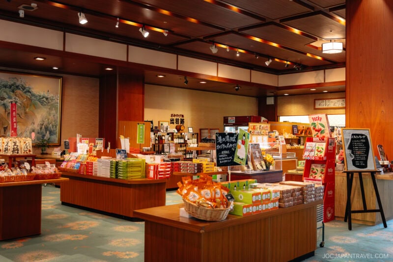 A well-lit Japanese gift shop in Yamagata features wooden shelves and tables displaying snacks, drinks, and souvenirs. Signs with Japanese text are visible around the store, and a large painting decorates one wall.
