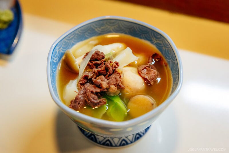 A bowl of Yamagata-style Japanese soup with thick noodles, sliced beef, vegetables, and fish balls in a light brown broth, served in a blue patterned bowl on a white surface.