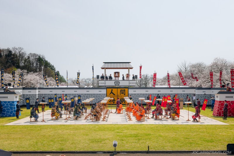 A large outdoor chessboard in Yamagata features people dressed as samurai warriors acting as chess pieces, with spectators watching from a raised platform and cherry blossom trees in the background.
