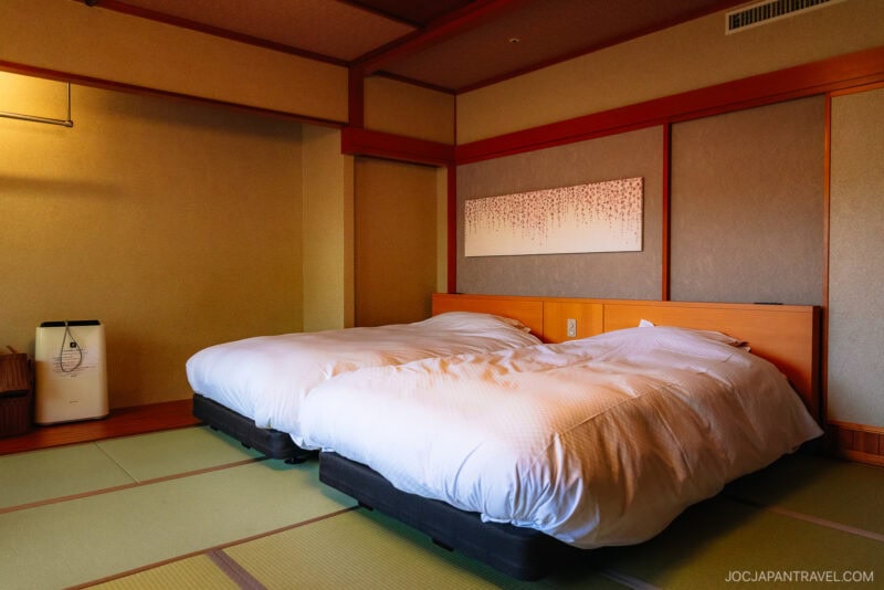 A traditional Japanese-style room in Yamagata with tatami mat flooring, two white beds, a wood-paneled wall, a modern air purifier, and decorative artwork above the headboard.