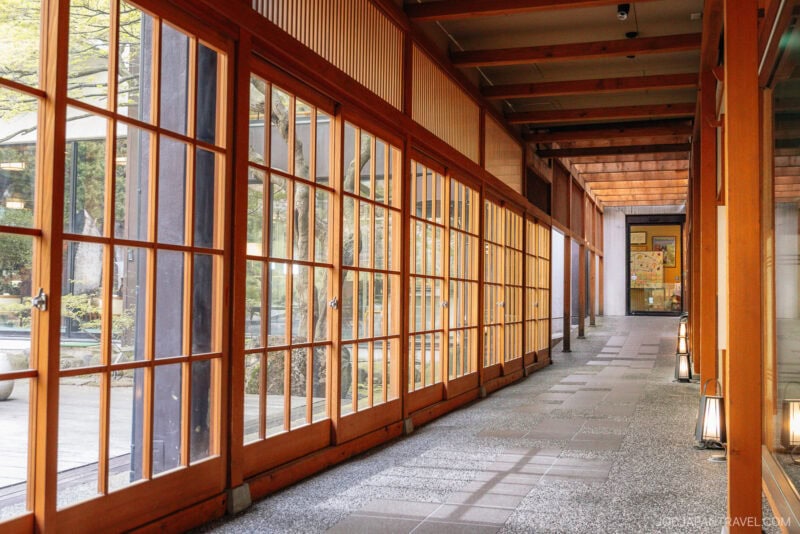 A corridor with wooden beams and large glass sliding doors on one side, letting in natural light. The floor is a mix of stone and tile, and small lanterns line the walkway, evoking serene yamagata design elements.