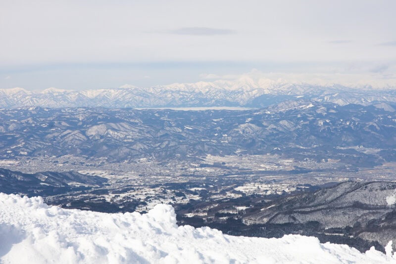 Snow-covered mountains and valleys stretch across the Yamagata landscape under a cloudy sky, with a layer of white snow in the foreground and distant peaks along the horizon.