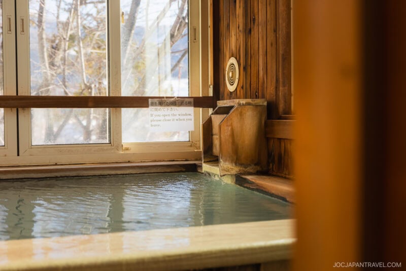 Indoor Japanese onsen bath with wooden walls and a window overlooking Yamagata’s lush trees. Water flows into the bath from a wooden spout, while a sign on the window provides instructions about closing it.
