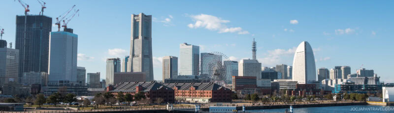 Panoramic view of the Yokohama city skyline with modern skyscrapers, a Ferris wheel, and the iconic sail-shaped InterContinental hotel by the waterfront under a clear blue sky.