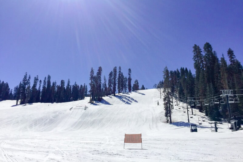 A bright, sunny day on a snowy ski slope in Yosemite National Park, lined with tall pine trees; an empty ski lift runs up the hill, and an orange safety barrier stands in the foreground.