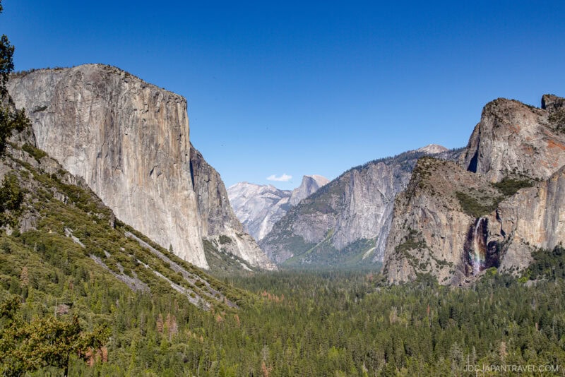 View of Yosemite Valley in Yosemite National Park featuring the granite cliffs of El Capitan on the left, Half Dome in the distance, and Bridalveil Fall on the right, with a forested valley floor beneath a clear blue sky.