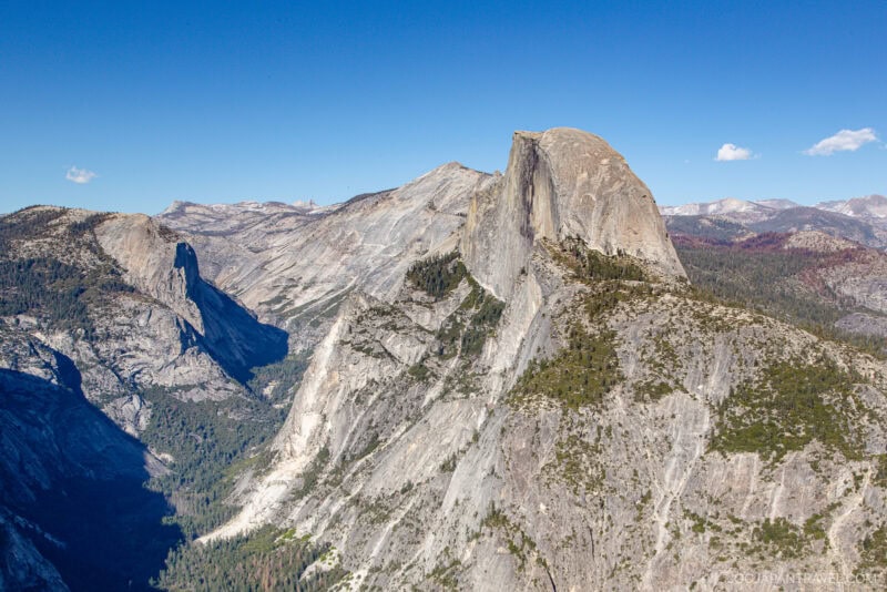 A clear view of Half Dome, a massive granite rock formation in Yosemite National Park, rises above forested valleys and rugged mountain terrain under a bright blue sky.