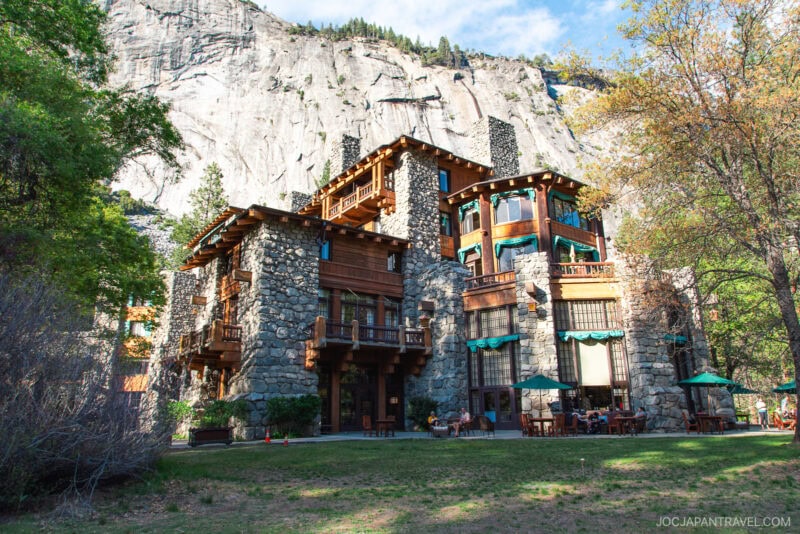 A large stone and wood lodge with balconies and green canopies sits at the base of a tall granite cliff in Yosemite National Park, surrounded by trees and a grassy lawn with outdoor seating.