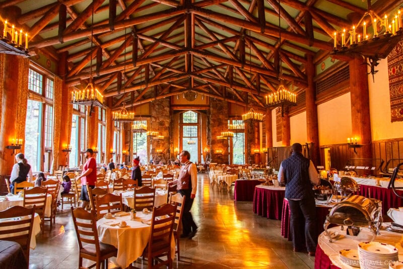 A grand dining hall in Yosemite National Park with a high wooden-beam ceiling, chandeliers, large windows, set tables, and people walking among the tables, creating a warm and inviting atmosphere.