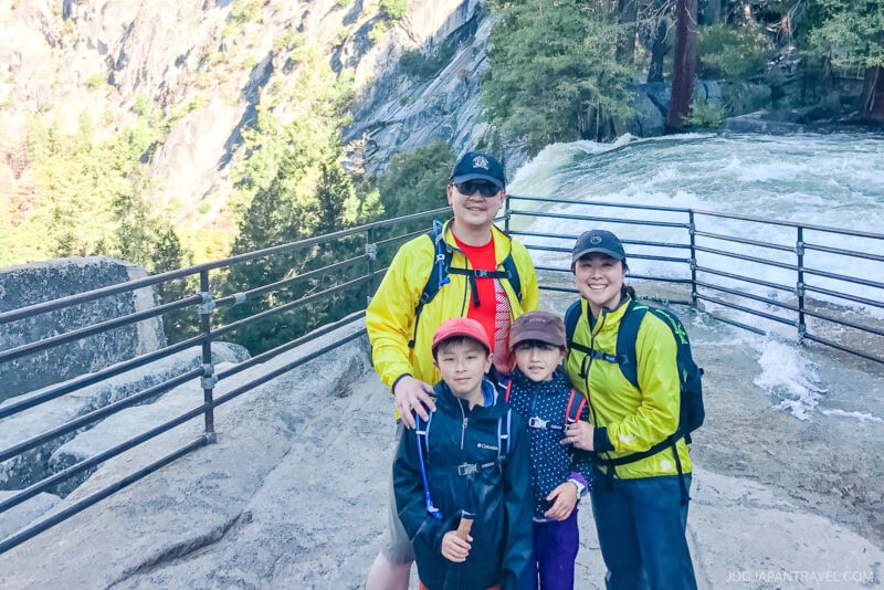 A family of four, dressed in outdoor clothing and backpacks, poses together near a metal railing by a river with rushing water and a forested, rocky background in Yosemite National Park.