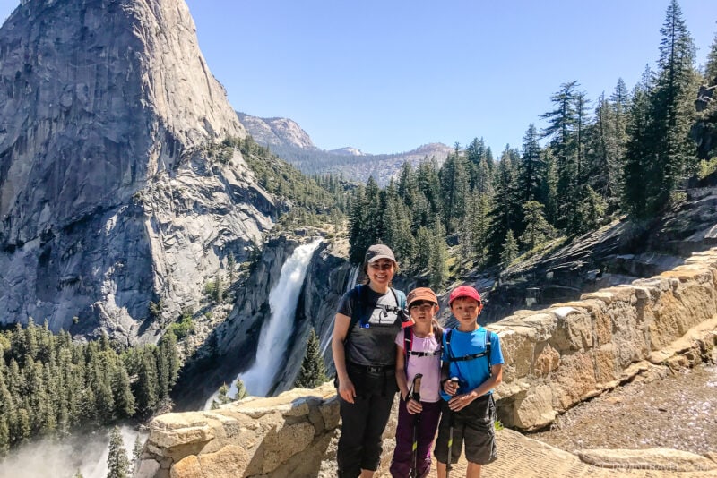 A woman and two children stand on a stone path in Yosemite National Park, with a scenic view of a waterfall, pine trees, and rocky mountains under a clear blue sky.