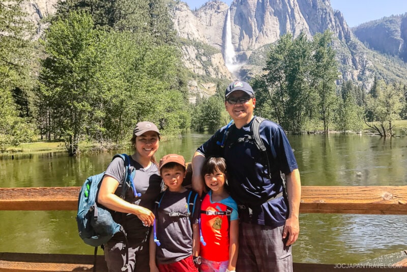 A family of four stands on a wooden bridge, smiling, with trees and a tall waterfall in the background on a sunny day at Yosemite National Park.