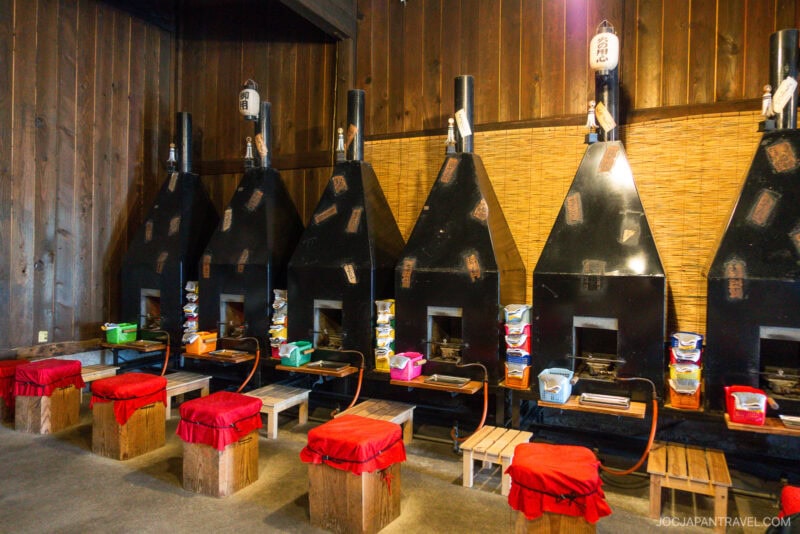 Five black, metal cooking stoves with individual seating and red-cushioned stools are lined up against a wooden wall at Yume Kojo Hida. Cooking utensils and ingredients are placed on each station.