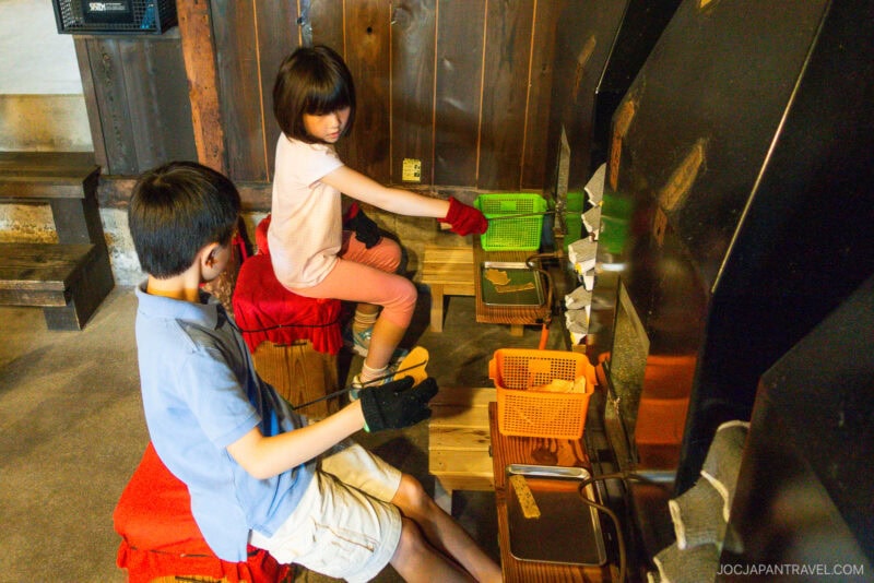 Two children sit on stools at Yume Kojo Hida, wearing gloves and holding metal tools and baskets as they make toast over a stone hearth indoors. Focused on cooking, theyre surrounded by wooden walls and benches.