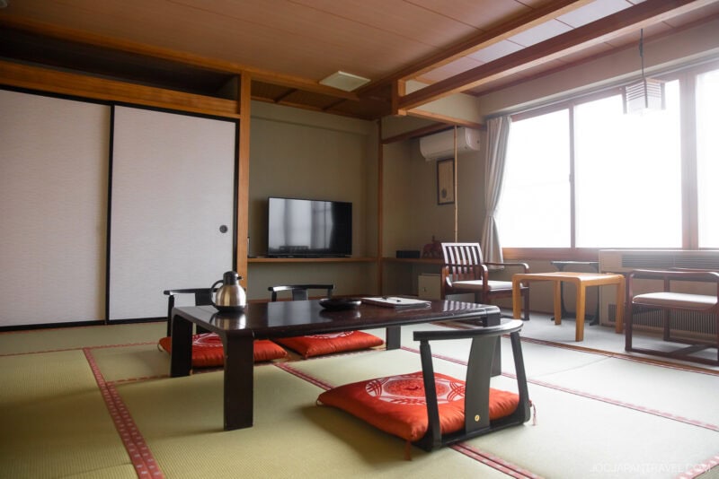 A traditional Japanese tatami room with a low wooden table, floor cushions, sliding shoji doors, and a window letting in natural light. Perfect for relaxing after exploring with your Zao winter guide; enjoy tea as snow falls outside.