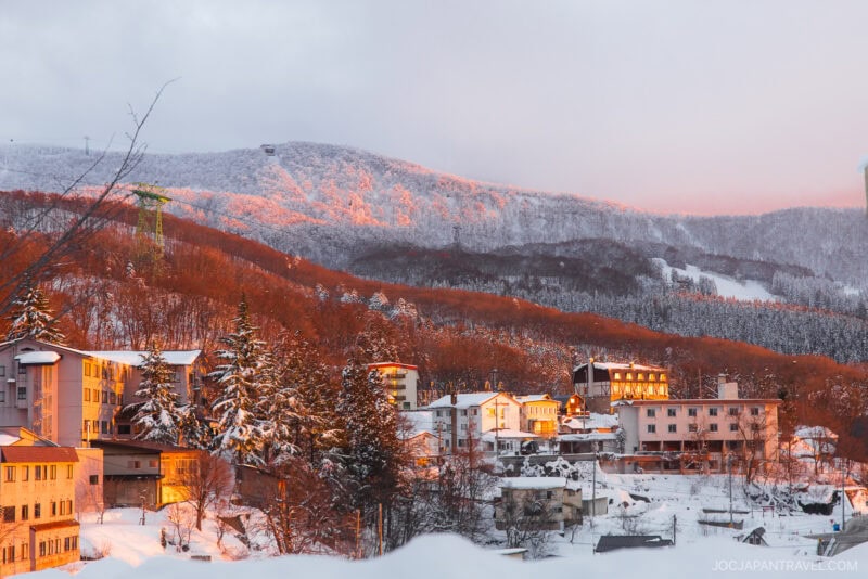 Snow-covered mountain town at sunset, with warm sunlight casting an orange glow on buildings and trees. In the background, forested hills and a cable car feature in this serene scene from the Zao winter guide under a partly cloudy sky.