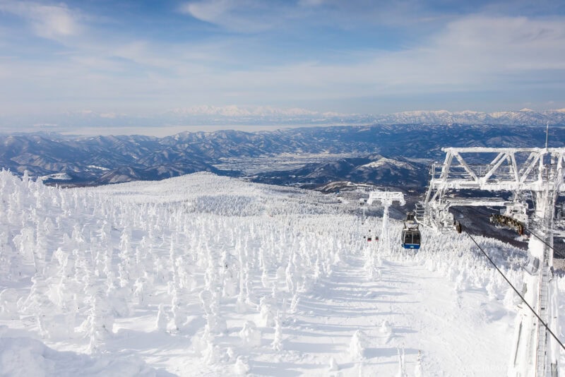 A snowy mountain landscape with snow-covered trees, a ski lift on the right, and distant mountain ranges under a blue sky with light clouds—perfect for your Zao winter guide adventure.