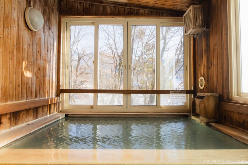 A wooden indoor hot spring bath with large windows overlooks snowy trees outside, letting in natural light and creating a warm, serene atmosphere—an ideal retreat featured in the Zao winter guide.