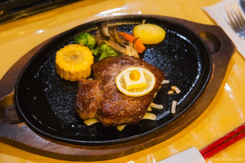 A grilled steak topped with a lemon slice and butter, served on a sizzling plate with corn on the cob, broccoli, potatoes, and carrots—a cozy meal perfect for your Zao winter guide adventures.