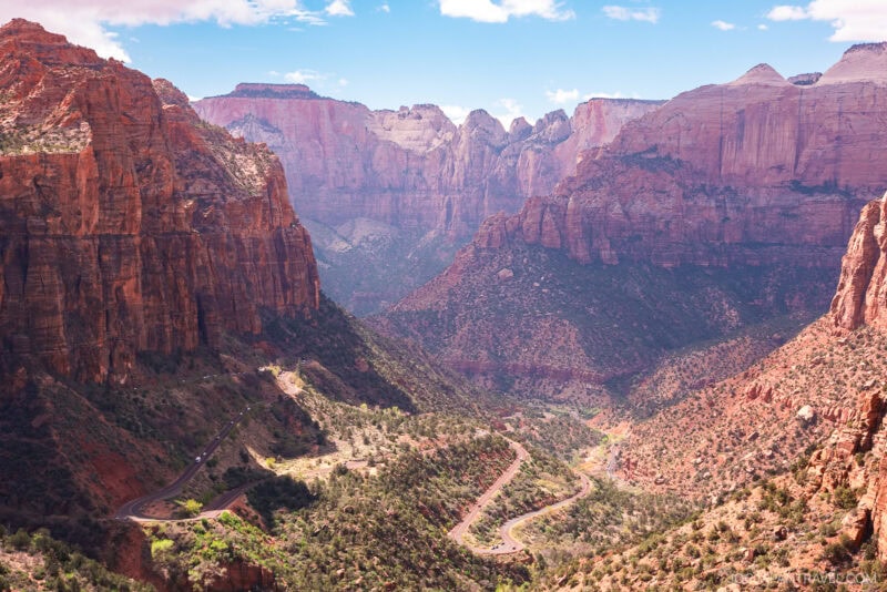 View of a winding road through a green valley surrounded by tall, dramatic red rock cliffs under a partly cloudy blue sky in stunning Zion National Park.