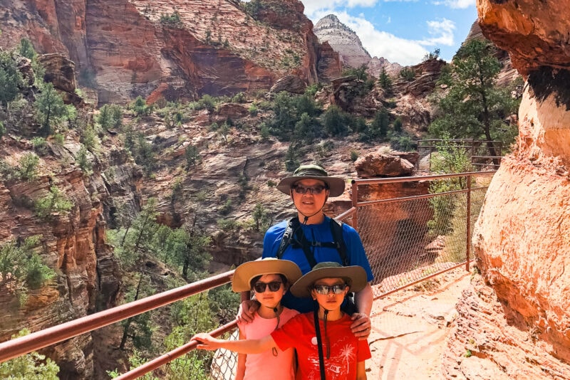 A man and two children wearing sun hats and sunglasses stand on a scenic Zion National Park canyon trail with red rock cliffs, trees, and a blue sky in the background. All are smiling and dressed for hiking.