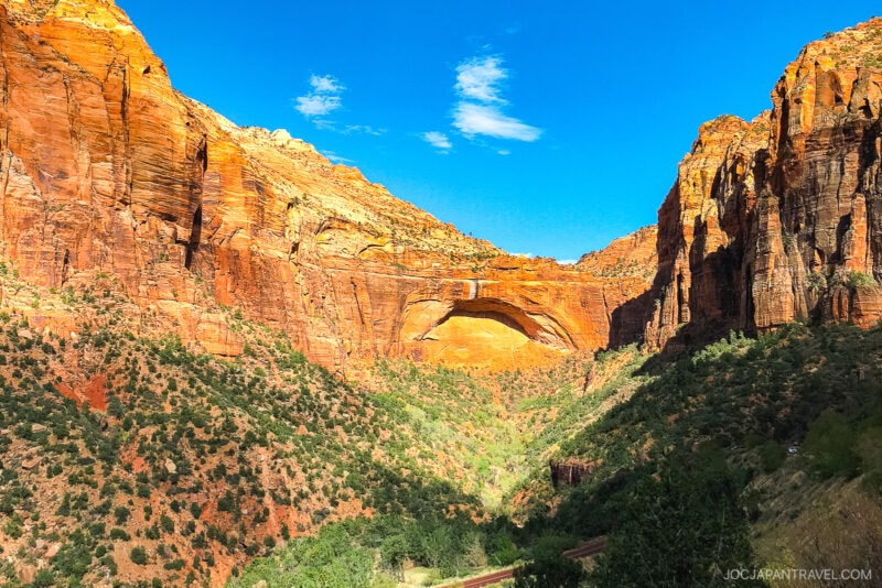 A vibrant canyon scene in Zion National Park with towering red rock cliffs, a natural arch in the center, and green vegetation scattered along the valley floor under a clear blue sky.