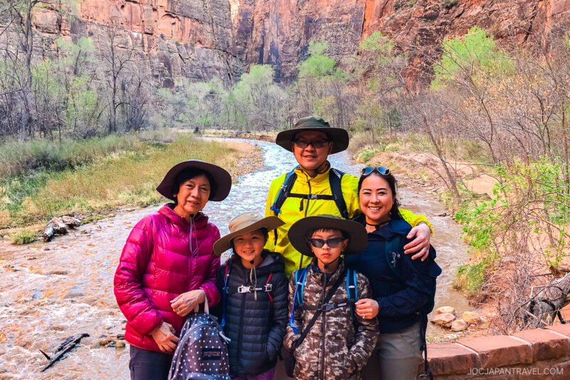 A family of five in hiking clothes and hats poses by a rocky river in a canyon with tall cliffs and sparse trees in Zion National Park. The group smiles, enjoying an outdoor adventure together.