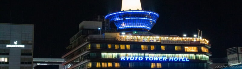 Night view of the illuminated Kyoto Tower Hotel, featuring blue and white lights, with the top of the Kyoto Tower visible above. Surrounding buildings are dimly lit.