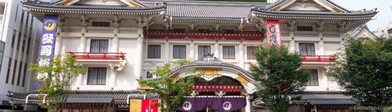 A traditional Japanese kabuki theater building with ornate architectural details, red and gold accents, and colorful banners hanging on both sides, partially obscured by green trees in the foreground.