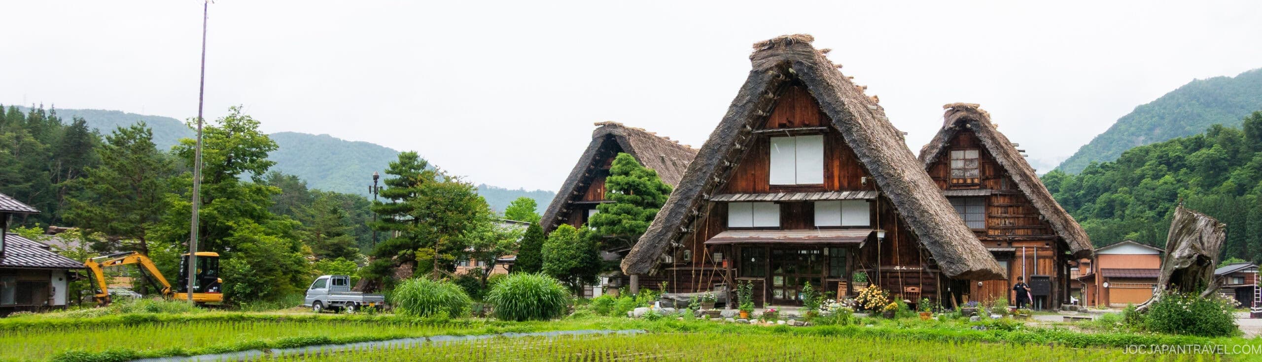 Traditional Japanese thatched-roof houses stand among lush greenery and rice fields, with mountains and cloudy skies in the background. Construction equipment and vehicles are visible to the left.