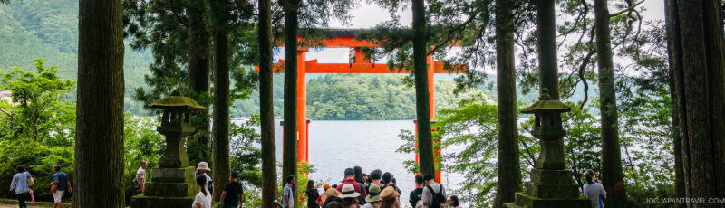 A group of people gather near a large, red torii gate by a lake, surrounded by tall trees and lush greenery. The scene is tranquil, with mountains visible in the background.