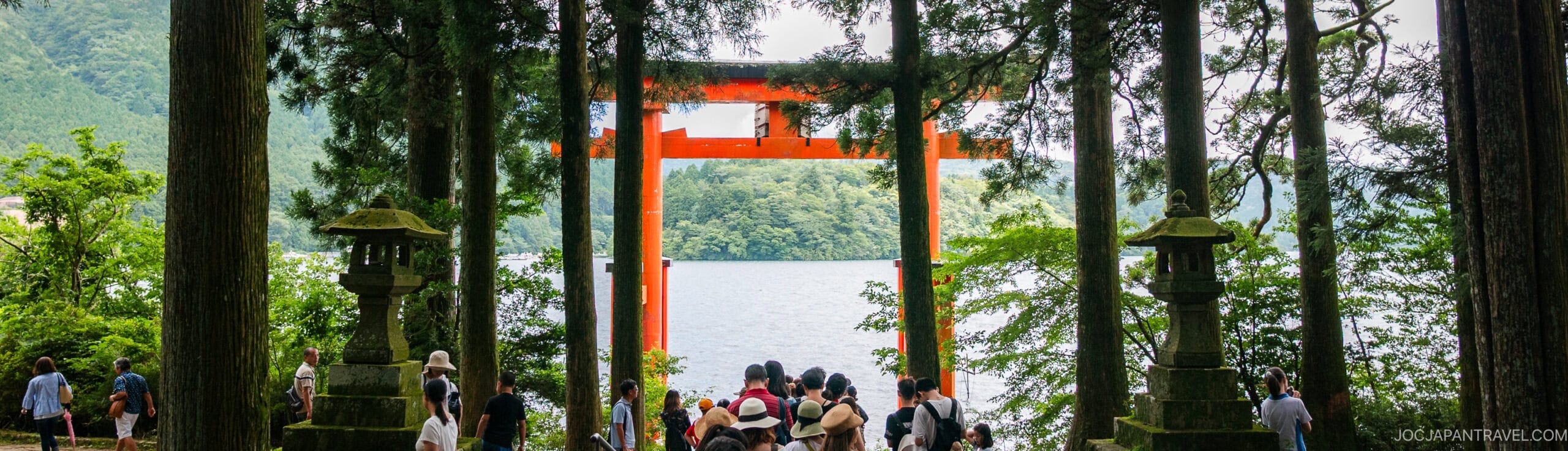 A group of people gather near a large, red torii gate by a lake, surrounded by tall trees and lush greenery. The scene is tranquil, with mountains visible in the background.