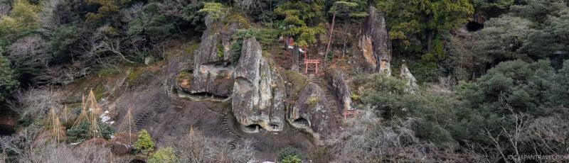 Large rocky outcrops surrounded by dense forest, with visible stairways and small red torii gates leading up the rocks, blending into the natural landscape.