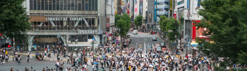 A large crowd of people crosses a busy intersection in an urban area, with tall buildings, shops, and a Starbucks Coffee in the background. Trees frame the scene on both sides.