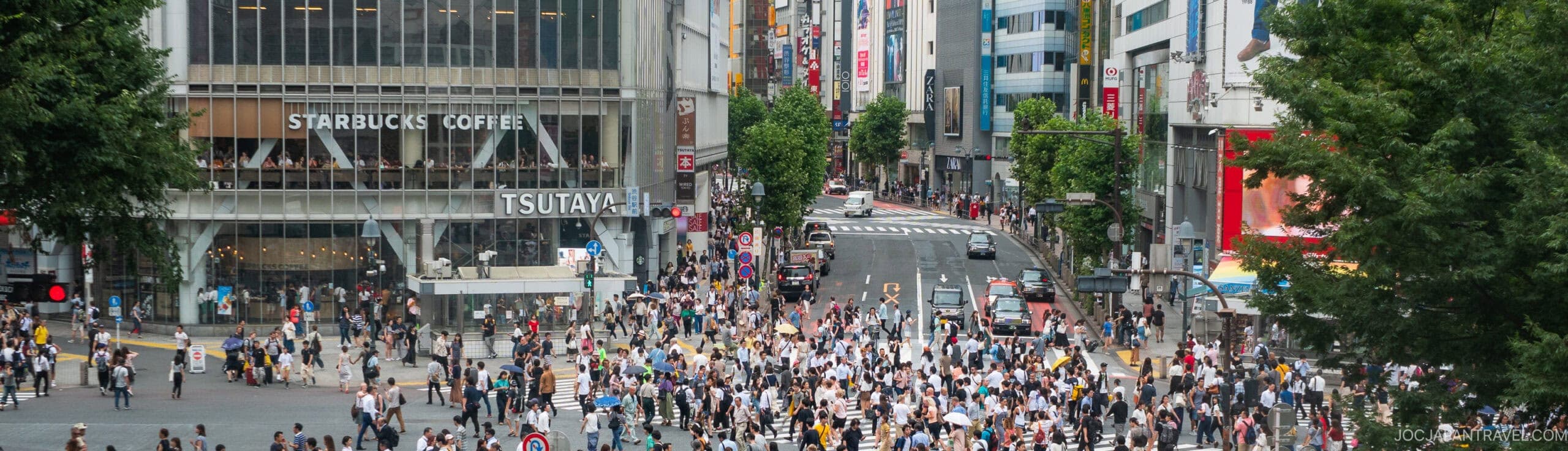 A large crowd of people crosses a busy intersection in an urban area, with tall buildings, shops, and a Starbucks Coffee in the background. Trees frame the scene on both sides.